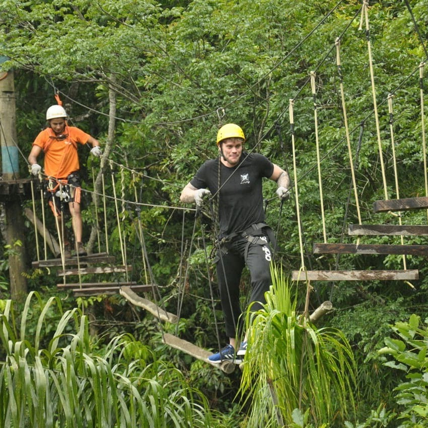 Dois homens praticando arvorismo em uma ponte suspensa de madeira em meio a uma floresta densa e verde.