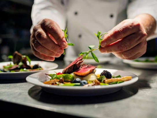 Chef finalizando prato gourmet com brotos verdes sobre carne selada, purê e frutas silvestres em ambiente de cozinha profissional.