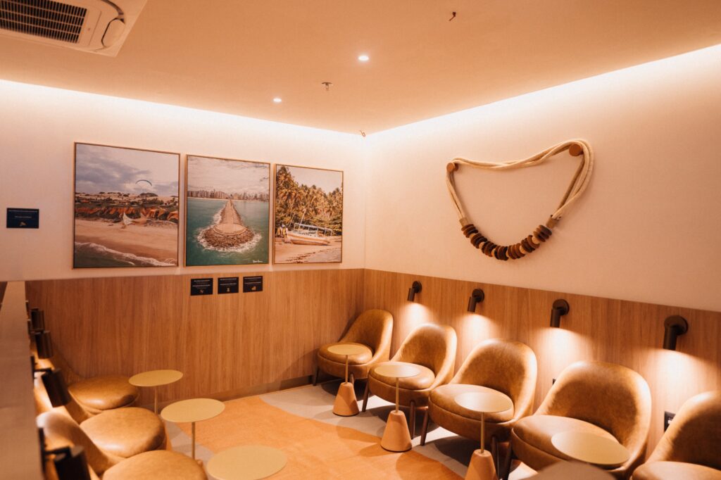 Sala de espera moderna com poltronas de couro, painéis de madeira, iluminação quente e três quadros de praia decorativos.